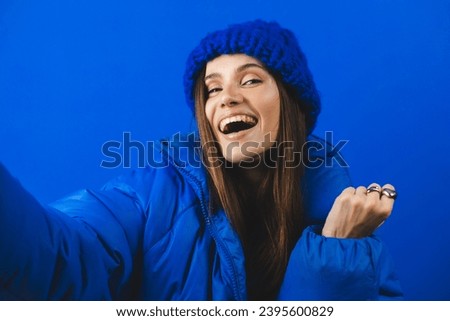 Shocked brunette woman in blue puffer jacket, hat talking speak on mobile phone isolated on blue background studio. Shocked woman with open mouth, hand near face wow win emotions. Girl make selfie.