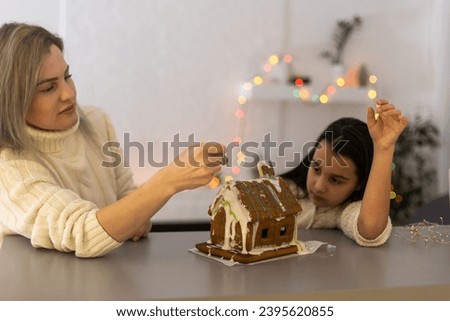 mother and daughter decorating gingerbread house. Beautiful living room with lights. Happy family celebrating holiday together.
