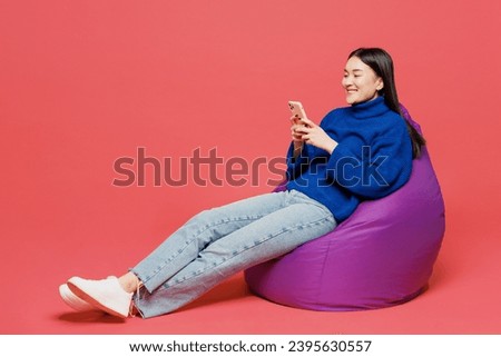 Full body young woman of Asian ethnicity she wearing blue sweater casual clothes sit in bag chair hold in hand use mobile cell phone isolated on plain pastel light pink background. Lifestyle concept
