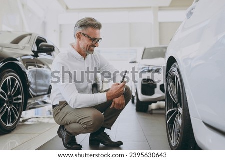 Adult man customer buyer client wear shirt take photo on mobile cell phone wheel rim tire choose auto want buy new automobile in car showroom vehicle salon dealership store motor show. Sales concept
