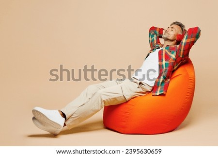 Full body adult man wear red shirt white t-shirt casual clothes sit in bag chair listen to music in headphones isolated on plain pastel light beige color background studio portrait. Lifestyle concept