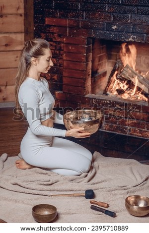 Delightful woman kneels on soft plaid and holds large metal musical bowl in her hands. Nearby lies a xylophone and other bowls. Fireplace background.