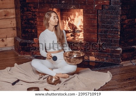 Girl with long hair sits on soft blanket with her legs folded and holding Tibetan bowl and xylophone in her hands. Closed eyes. Fireplace background.