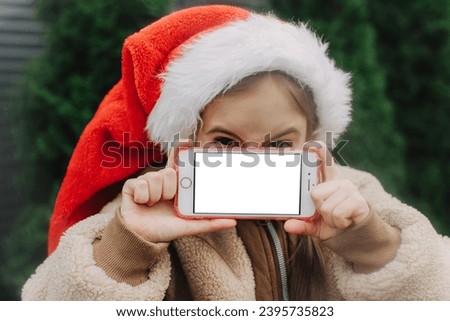 Little girl in Christmas Santa hat shows phone with white screen. Chroma key white screen smartphone set up for advertising. Cute child looking at camera. Adorable kid with digital device. Copy space.
