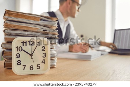 Close up clock and stack of red tape paper overload on office table, with punctual productive efficient company employee working with data on laptop in background. Business, time, paperwork concept
