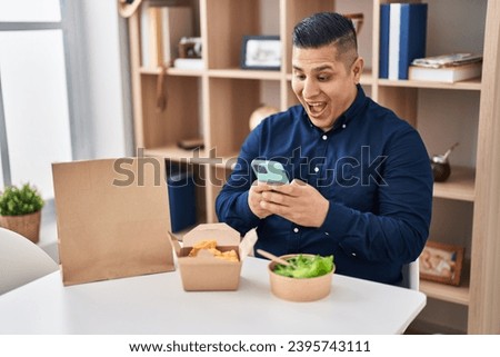 Hispanic young man eating take away food using smartphone smiling and laughing hard out loud because funny crazy joke. 