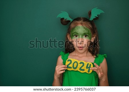 A surprised girl dressed up as a green dragon, holding the figures of the new year 2024 in her hands. A symbol according to the Eastern calendar. Dinosaur wings made of paper on a child's head