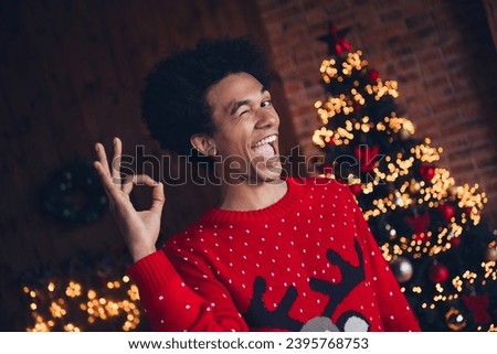 Photo of cool funny guy dressed red ugly x-mas pullover blinking showing okey gesture celebrating new year indoors house room