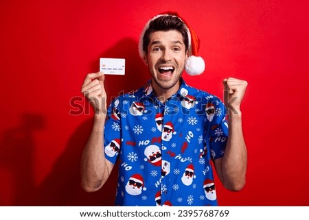 Photo of crazy guy wear christmas hat print shirt clenching fist demonstrate credit card get salary isolated on vivid red color background