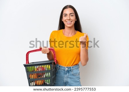 Young caucasian woman holding a shopping basket full of food isolated on white background making money gesture