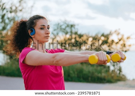 American athlete trains with dumbbells outdoors listens to music using sports headphones looks into the distance and dreams of winning the Olympics Royalty-Free Stock Photo #2395781871