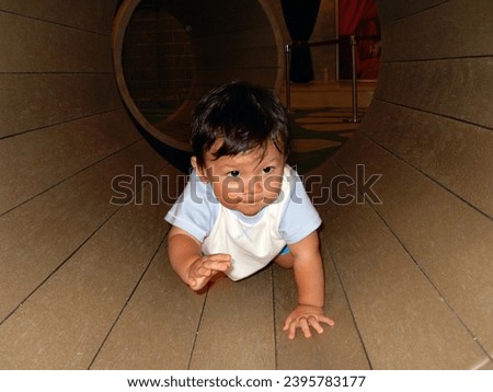 interior photo view of a handsome good looking eurasian cute chinese baby toddler crawling inside a large big toy barrel tunnel cave for kid child children in a kindergarten playground 