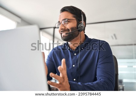 Happy Indian call center agent wearing headset talking to client working in customer support office. Professional contract service telemarketing operator using laptop having conversation. Candid shot Royalty-Free Stock Photo #2395784445