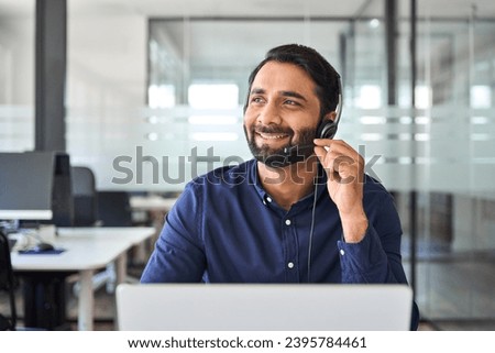 Smiling happy Indian call center agent wearing headset talking to client, contract service telemarketing operator using laptop having conversation working in customer tech assistance support office. Royalty-Free Stock Photo #2395784461