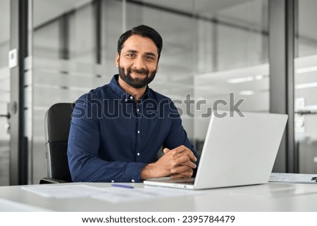 Smiling confident Indian business man employee looking at camera sitting at work desk with laptop computer. Portrait of smart happy businessman office worker or entrepreneur posing at modern workplace Royalty-Free Stock Photo #2395784479