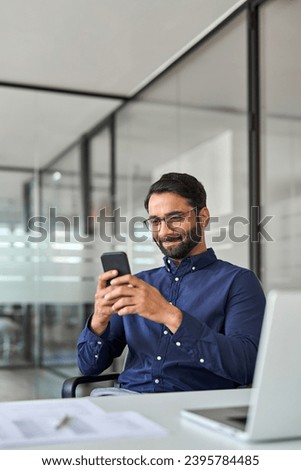 Professional Indian business man working using smartphone in office. Vertical shot. Smiling company employee, busy businessman holding cellphone looking at mobile phone sitting at workplace desk. Royalty-Free Stock Photo #2395784485