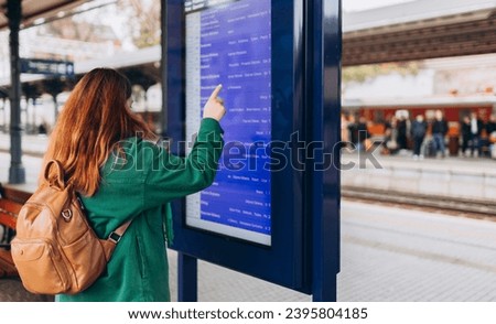 Young redhead woman with backpack pointing finger on train timetable on a railway station. 30s female with smart phone Looking at Arrival and Departure Information Display, Traveler Royalty-Free Stock Photo #2395804185