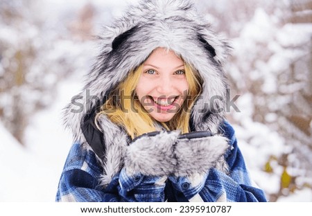 Wintertime. Closeup portrait of smiling winter girl in snowy park. Winter holidays and vacation. Beautiful woman in warm plaid coat, fur hat and mittens. Winter fashion for women. Cold winter weather.