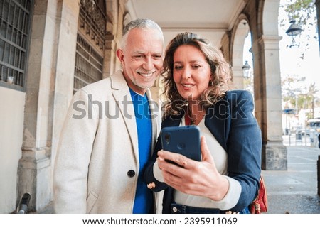 Mature tourists couple using a smartphone and watching funny pictures on their weekend trip and enjoying together. Middle aged woman and her husband searching hotel on a cell phone standing outside