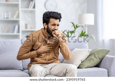 Indian young man sitting at home on the couch and coughing, covering his mouth, holding his chest, feeling severe pain, suffering from an infectious disease. Royalty-Free Stock Photo #2395919765