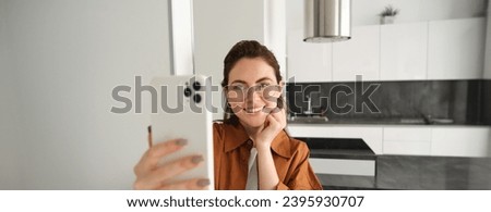 Beautiful young woman in glasses taking selfie in her flat, using smartphone, posing for photo, standing with mobile phone and smiling.