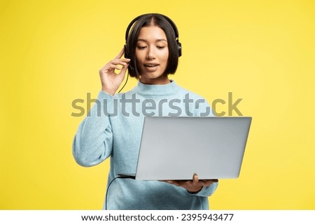 Attractive beautiful Asian businesswoman, saleswoman holding laptop, wearing headphones, talking, answering call standing isolated on yellow background. Contact center concept Royalty-Free Stock Photo #2395943477