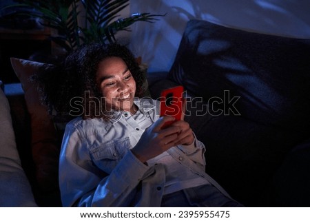 Joyful young black girl using cell phone lying on sofa. Nice afro woman enjoying chatting on mobile at night home. Single African American female generation z people on dating apps and social networks Royalty-Free Stock Photo #2395955475