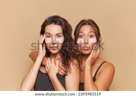 Two pretty young women stand on beige background hugging, both with patches under their eyes, skincare concept, copy space.