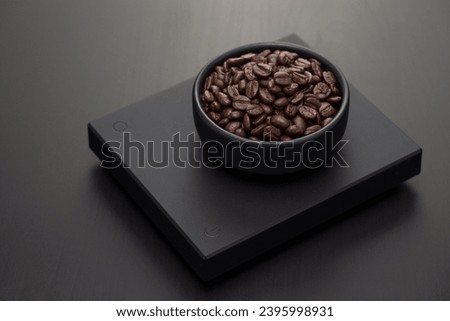 Dark roasted coffee beans a small black ceramic bowl on a black digital scale.