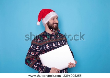 Bearded man in knitted sweater red Christmas Santa Claus hat hold in hand closed laptop pc computer isolated on blue background. Happy New Year 2024 celebration. 