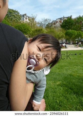 exterior photo view of a chinese mother view from the back in a parisian paris public park garden holding her baby toddler eurasian handsome good looking male boy who is looking at the photographer