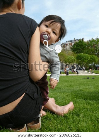 exterior photo view of a chinese mother view from the back in a parisian paris public park garden holding her baby toddler eurasian handsome good looking male boy who is looking at the photographer