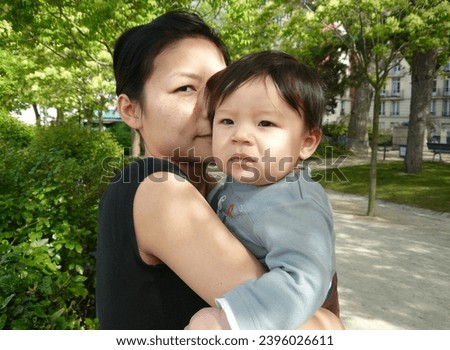 exterior photo view of a young beautiful charing pretty chinese mother woman girl view in a parisian paris public park garden holding her baby toddler eurasian handsome good looking male boy family