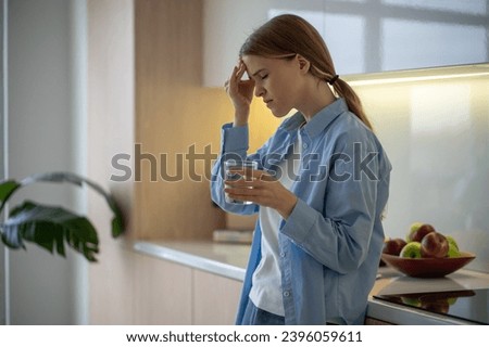 Woman feeling terrible pain in head drinking water on kitchen at home with closed eyes. Girl having headache, migraine, cephalalgia, premenstrual syndrome. Taking medications, painkillers concept. Royalty-Free Stock Photo #2396059611