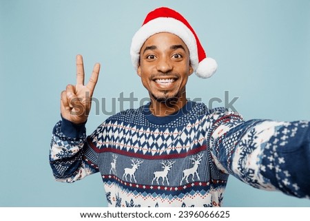 Close up young man wears sweater Santa hat posing doing selfie shot pov on mobile cell phone show v-sign isolated on plain blue background. Happy New Year 2024 celebration Christmas holiday concept