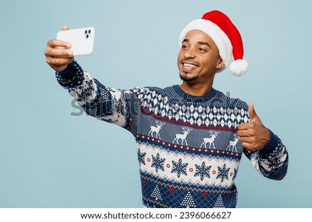 Young man wear knitted sweater Santa hat posing do selfie shot on mobile cell phone show thumb up isolated on plain pastel blue background. Happy New Year 2024 celebration Christmas holiday concept