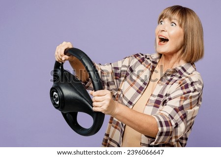 Side view elderly excited happy blonde woman 50s years old wear beige t-shirt shirt casual clothes hold steering wheel driving car isolated on plain pastel light purple background. Lifestyle concept