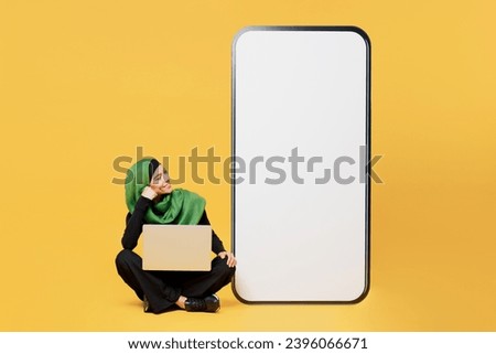 Full body young asian muslim woman wear green hijab abaya black clothes big huge blank screen mobile cell phone use laptop pc isolated on plain yellow background. People uae islam religious concept