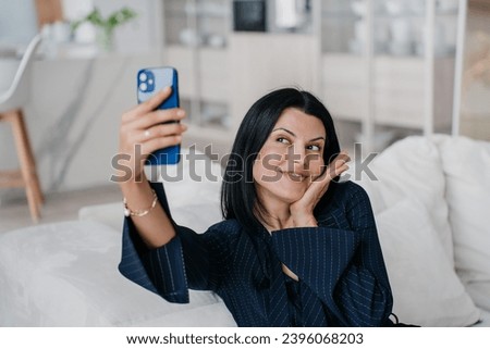 cheerful American brunette polygamous dress is sitting on a comfortable sofa at home taking selfies using her phone, smiling with her health and physical appearance. Successful people