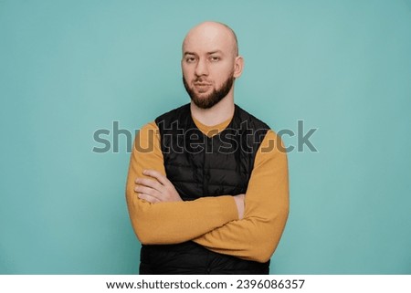 Bald caucasian bearded man in casual clothes standing hands folded on chest looks at camera with focused calm face against turquoise studio background with copy space. Royalty-Free Stock Photo #2396086357