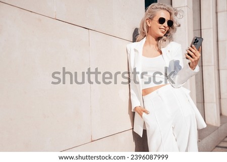 Young beautiful blond woman wearing nice trendy white suit jacket. Smiling model posing in street. Fashionable female outdoors. Cheerful and happy. Holds smartphone, uses phone apps, looks at screen Royalty-Free Stock Photo #2396087999