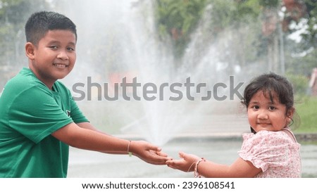 Asian brother and sister showing affection between family members, older sister in a green t-shirt reaching out her hand to her younger sister with a fountain in the playground in the background