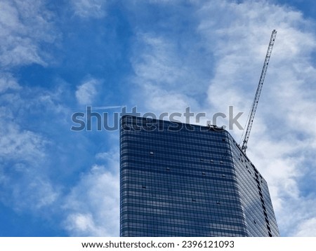 Modern skyscraper with a reflective glass facade under a blue sky scattered with white clouds. A crane is attached to the building, suggesting ongoing construction or maintenance