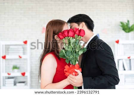 Asian couple lover romantic kissing behind the red roses bouquet after give flower for dating in Anniversary day while young husband wearing silver wedding ring and holding wife hand together