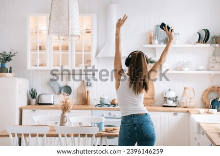 rear view of a positive girl in jeans dancing in the kitchen at home using headphones and a phone that raised her hands up shows a victory sign a fun weekend at home