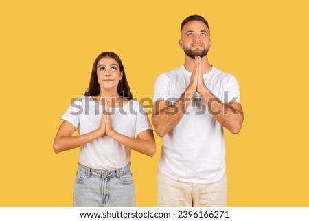 Positive millennial european family in white t-shirts hoping or wishing and make pray gesture, look up at free space, isolated on yellow studio background. Lifestyle, dream come true