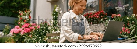 Beautiful flower shop owner wearing apron working on laptop in her store Royalty-Free Stock Photo #2396173929