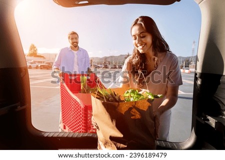 Young arab woman joyfully unpacks fresh groceries from paper bag in her car's trunk as happy man approaches with a cart on background, view from inside auto Royalty-Free Stock Photo #2396189479