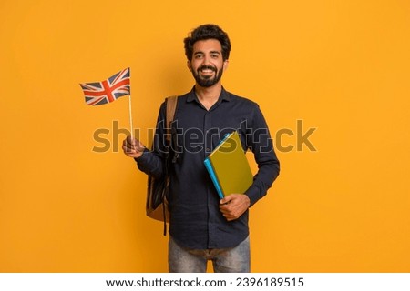 Language Courses. Smiling Young Indian Man Holding British Flag And Workbooks While Standing Over Yellow Background In Studio, Eastern Man Recommending Remote Education Programs, Copy Space Royalty-Free Stock Photo #2396189515