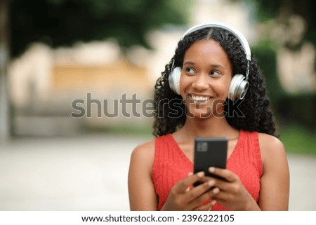Front view portrait of a happy black woman looks at side listening to music walking in the street Royalty-Free Stock Photo #2396222105